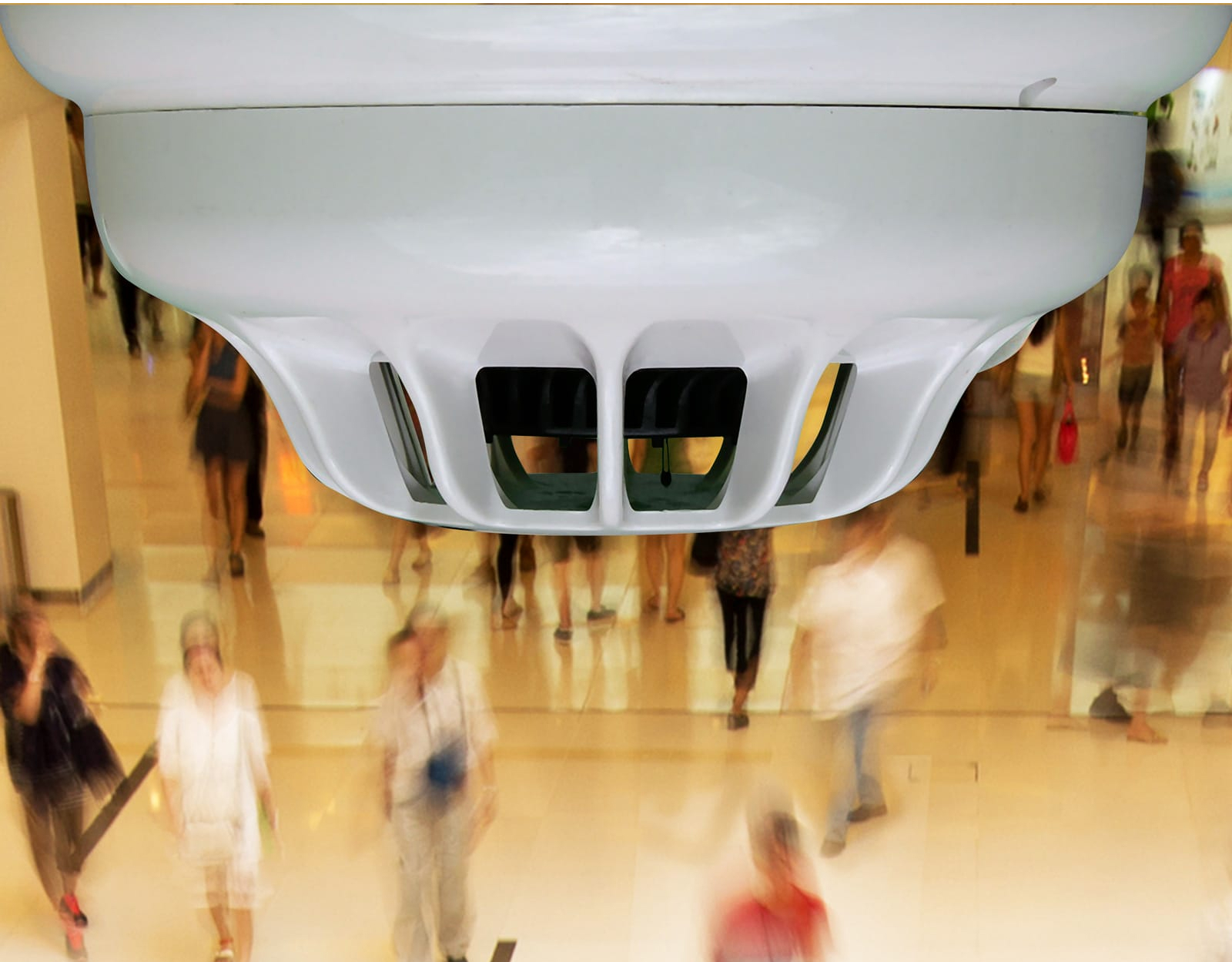 Close-up of a fire detection unit on a ceiling in the foreground, with people walking below