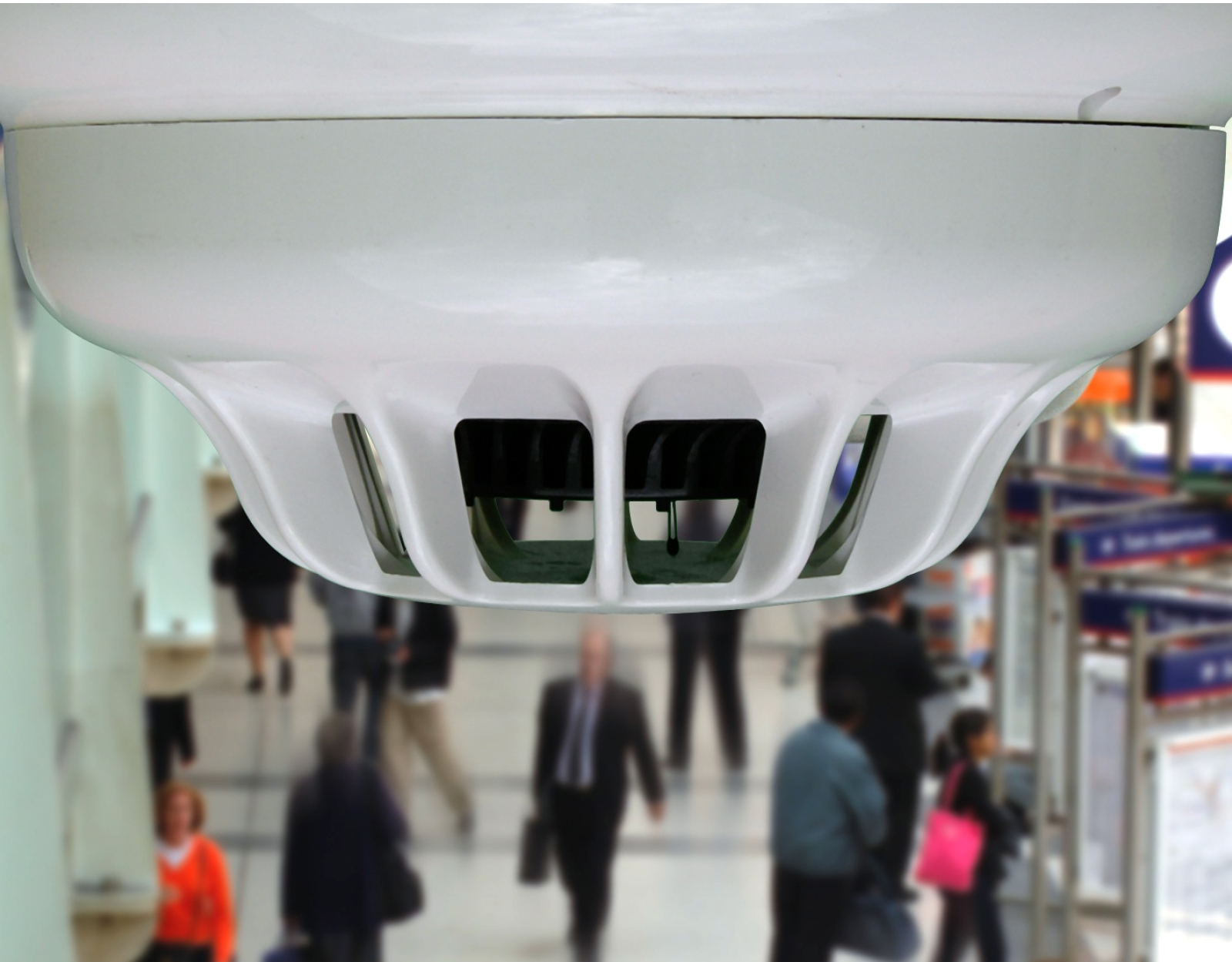 Close-up of a fire detection unit mounted on a ceiling, with people walking below in the background