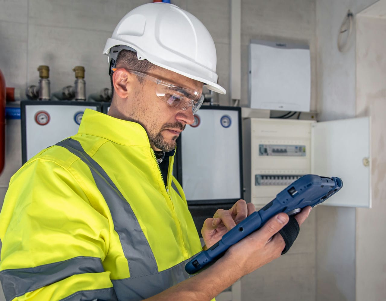 An electrical technician reading a tablet while standing near a switchboard with fuses.